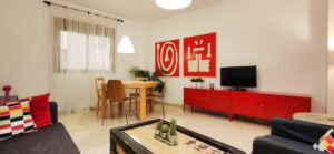 Salón con mueble de TV rojo, sofá y butacón oscuros. Mesa redonda con 4 sillas y mesa de centro.