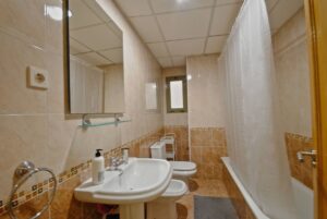 Baño con bañera a la derecha con cortina, y a la izquierda lavabo con espejo, bidé, inodoro y carrito blanco. Ventana pequeña al fondo.