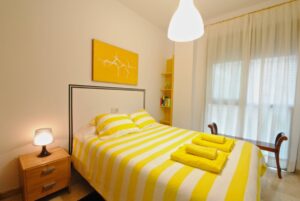 Dormitorio con cama doble decorada con cojines y toallas, mesita de noche, estantería, banco para maletas, y ventanal hasta el suelo.