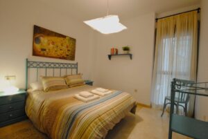 Dormitorio con cama doble decorada con cojines y toallas, 2 mesitas de noche, silla y ventana hasta el suelo.